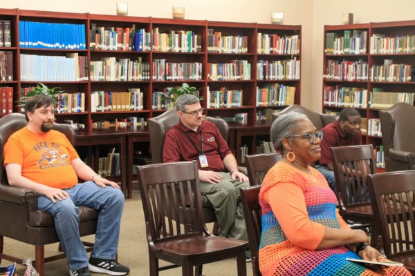 Four adults sit spaced out on wooden chairs in a library, facing forward. Bookshelves filled with books line the wall behind them. The atmosphere appears calm and attentive.