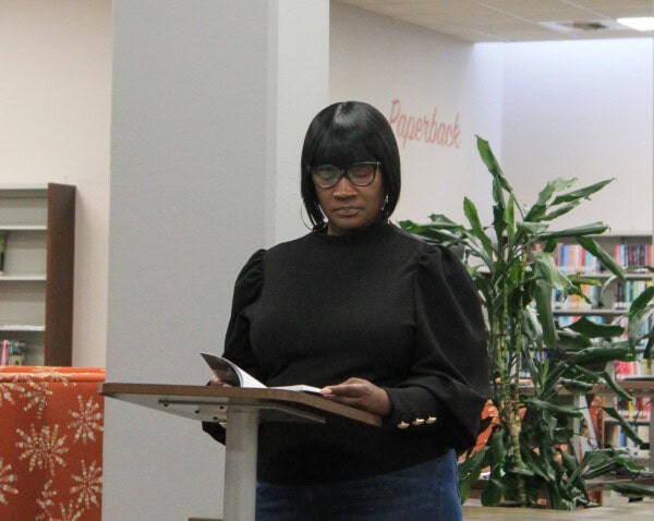 A woman wearing glasses and a black top stands at a podium reading from a book in a library, with bookshelves and a plant visible in the background.