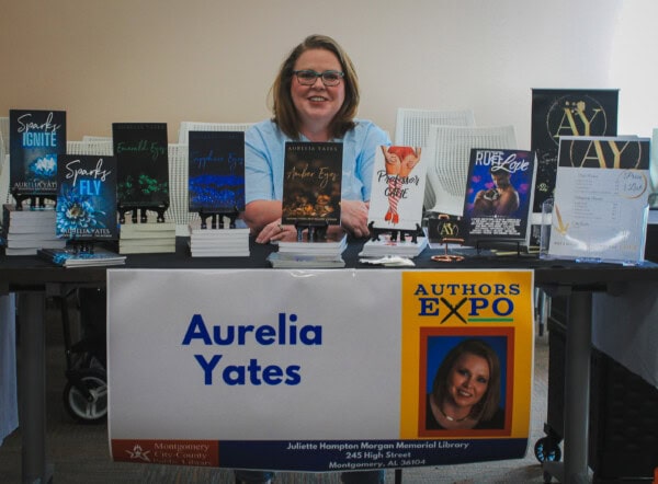 A smiling woman sits behind a table displaying her books at an Authors Expo. A banner reads Aurelia Yates with her photo and library information. Various book covers and promotional materials are arranged on the table.