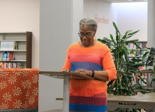 A woman wearing glasses and a colorful orange and blue dress stands at a podium, reading or speaking in a library with bookshelves and green plants in the background.