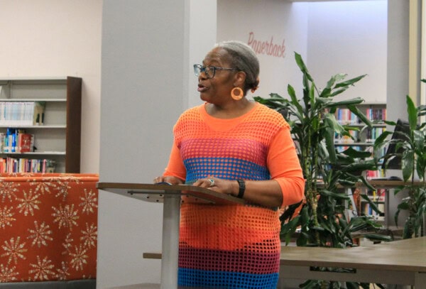 An older woman wearing glasses and a colorful orange and blue dress speaks at a podium in a library, with bookshelves and green plants in the background.