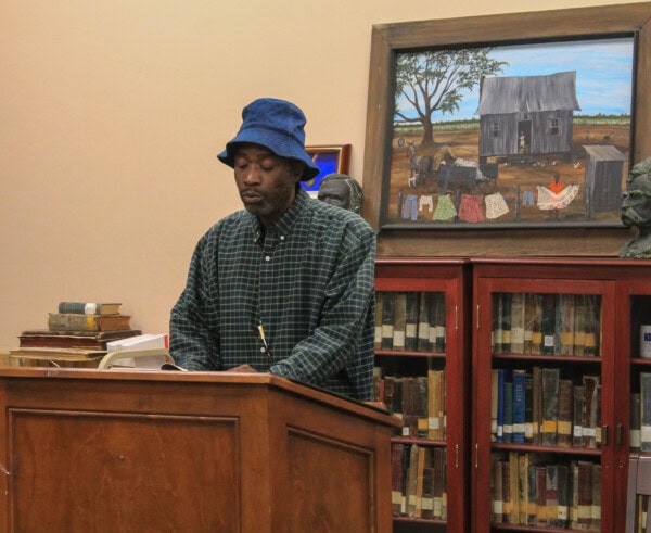 A man in a blue bucket hat and plaid shirt stands at a wooden podium in a library, with bookshelves, stacked books, and a painting of a rural scene visible in the background.