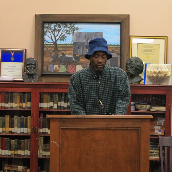 A man in a blue hat reads at a podium in a library, surrounded by bookshelves, busts, framed awards, and artwork featuring a house in a rural landscape.