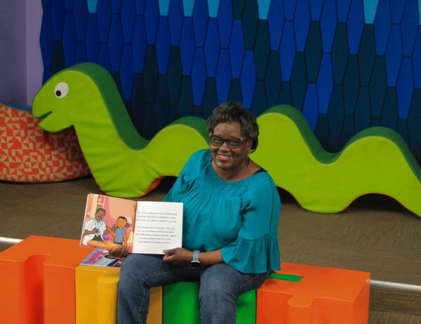 A smiling woman wearing glasses and a teal top sits on colorful foam blocks, holding open a children’s book. Behind her is a large green caterpillar-shaped seat and a blue-patterned wall.