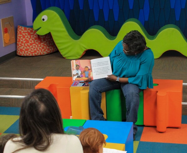 A woman sits on colorful benches reading a picture book to a small group of children in a playroom with a large green caterpillar decoration in the background.