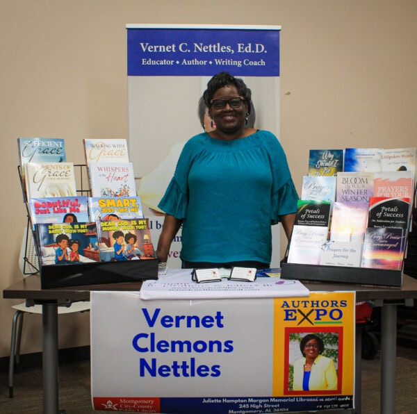 A woman in a teal blouse stands smiling behind a table displaying her books at an expo. A sign on the table reads Vernet Clemons Nettles, and various books are arranged upright for viewing.
