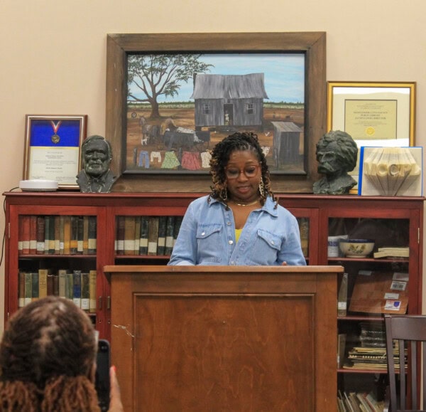 A woman with braided hair and glasses stands at a podium, reading or speaking in a library. Behind her are bookshelves, framed art, and two small bust sculptures. An audience member is visible in the foreground.
