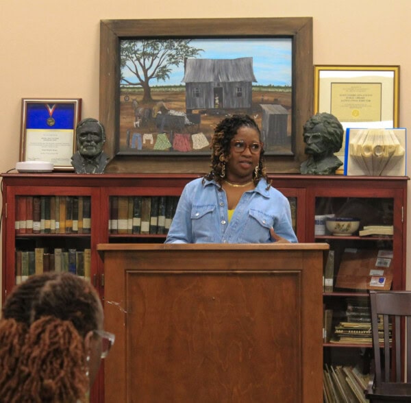 A woman with glasses stands at a wooden podium, speaking in front of a bookshelf with books, framed pictures, awards, and two bust sculptures. A rustic house painting is displayed above the bookshelf.