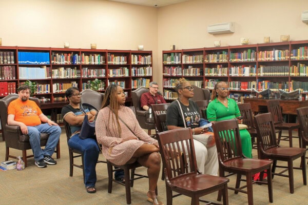 A small group of adults sit spaced apart on chairs in a library, attentively facing forward. Bookshelves line the walls, and there are books and bags next to some attendees.
