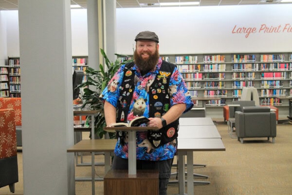 A man with a beard, wearing a colorful shirt and patch-covered vest, stands at a podium with an open book in a library. Shelves of books and a sign reading Large Print F are visible in the background.