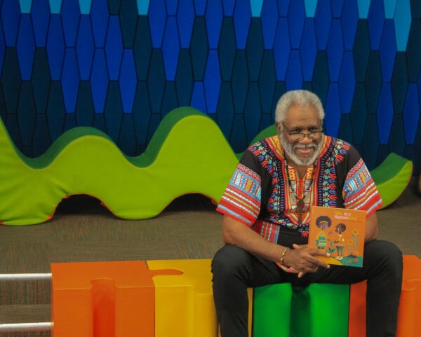 An older man with gray hair and glasses sits on a colorful bench, smiling and holding a childrens book. He wears a vibrant, patterned shirt. Behind him is a blue and green wavy backdrop.
