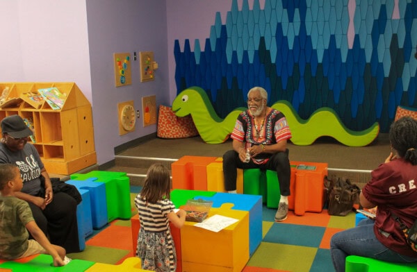 A man sits on a green block reading to children in a colorful playroom with a green snake decoration on the wall. Adults and children sit or stand on colorful foam puzzle pieces, listening attentively.