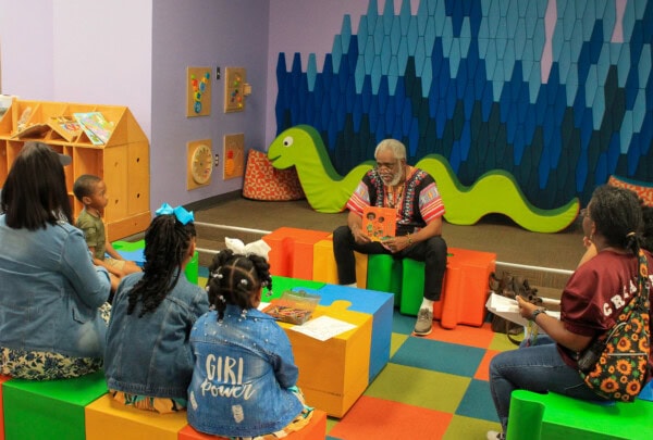 A man reads a book to a group of children and adults in a colorful library room with a green snake decoration and puzzles on the floor. The children sit on colorful cube seats, listening attentively.