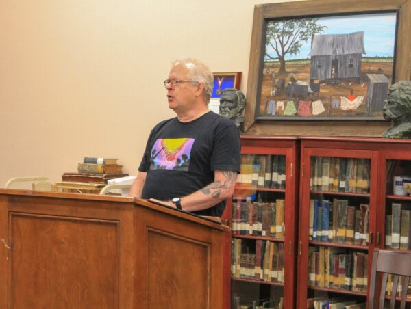 A man with glasses and a tattoo, wearing a graphic t-shirt, speaks at a wooden podium in a library. Behind him are bookshelves with books, bust sculptures, and a painting of a rural scene with a house and laundry.