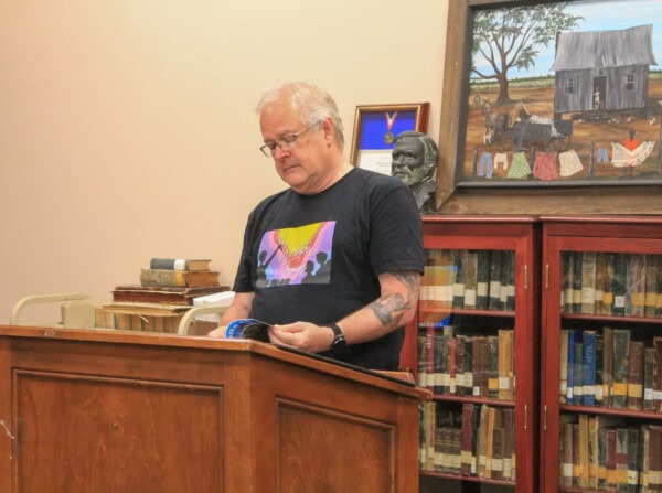 A man with gray hair and glasses stands at a wooden podium, reading from a book in a library. Behind him are bookshelves, a framed portrait, a painting, and a sculpture. He wears a black t-shirt with a colorful graphic.