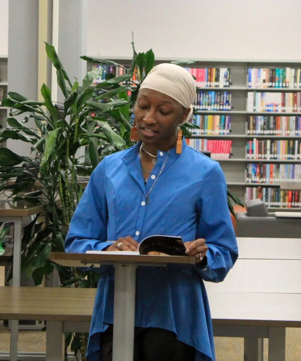 A person wearing a blue shirt and a cream headscarf reads from a book at a podium in a library, with bookshelves and a large plant in the background.