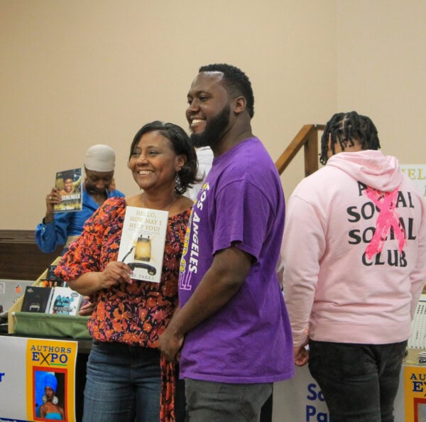 A smiling woman and man pose together at a book fair, with the woman holding a book titled Hello, How May I Help You? Other people browse books in the background.