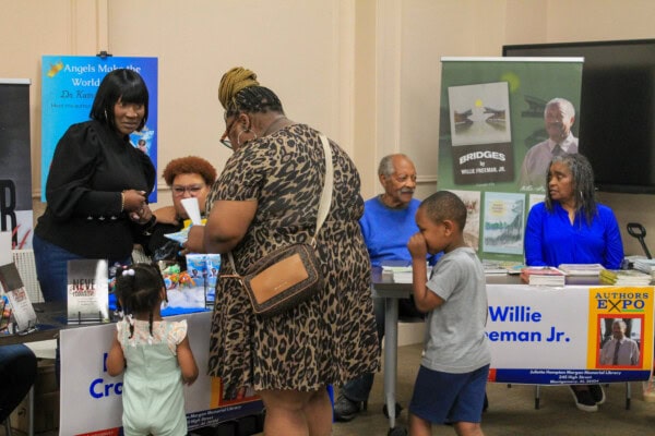 Several adults and children interact at a book fair or expo, standing at tables displaying books, posters, and signs featuring authors and their works. The atmosphere appears friendly and engaging.