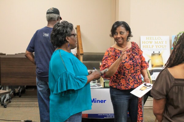 Two women stand and converse at an indoor event; one holds a book and gestures. A man and another person are seen from behind. A sign in the background reads, HELLO, HOW MAY I HELP YOU?.