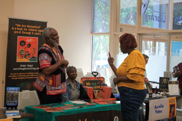 A man in a colorful shirt speaks to a woman in yellow at a book fair table displaying children’s books, including “We Are Humankind.” Another woman sits behind the table. Book displays and posters are visible in the background.