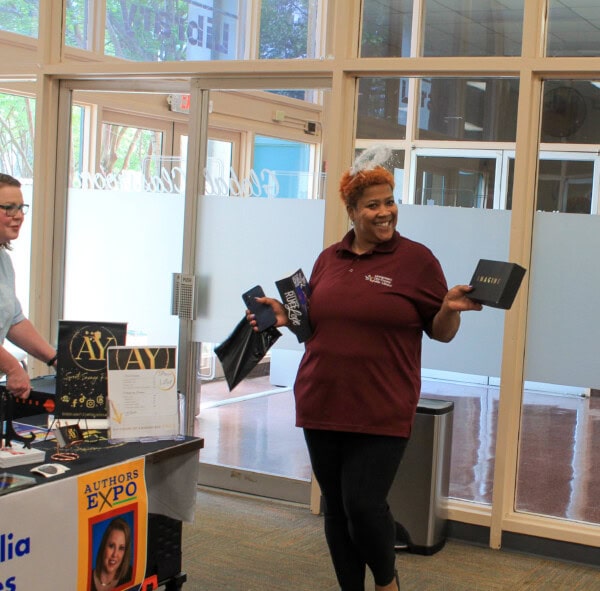 A smiling woman in a maroon polo shirt holds up a book and a black bag while standing near a table with books and an Authors Expo sign, inside a bright room with large windows.