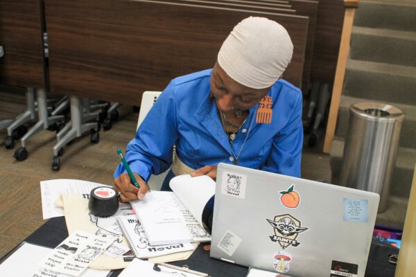 A person wearing a blue shirt and white headscarf writes at a table covered with papers, zines, and a laptop decorated with various stickers. The setting appears to be an indoor workspace or classroom.