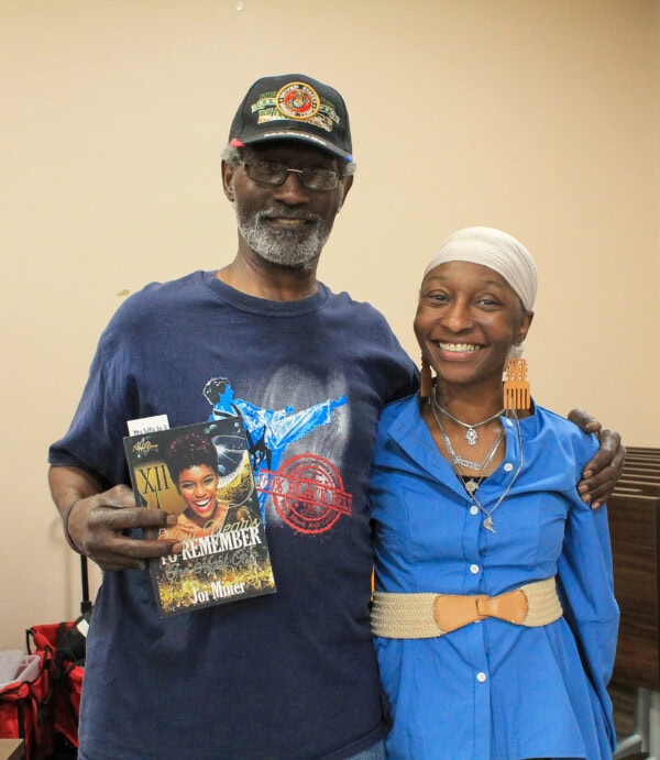 A smiling man in a military cap holds a book while standing with his arm around a smiling woman in a blue dress and headscarf. The background is an indoor setting with neutral-colored walls.