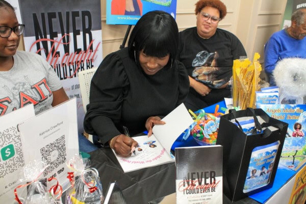 A woman in black signs a colorful children’s book at a table covered with books, gift bags, and promotional materials, while two people sit nearby and watch.