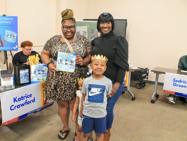 Two women and a smiling boy wearing a crown pose together at a book event. One woman holds a children’s book. Tables with books and promotional signs are visible in the background.