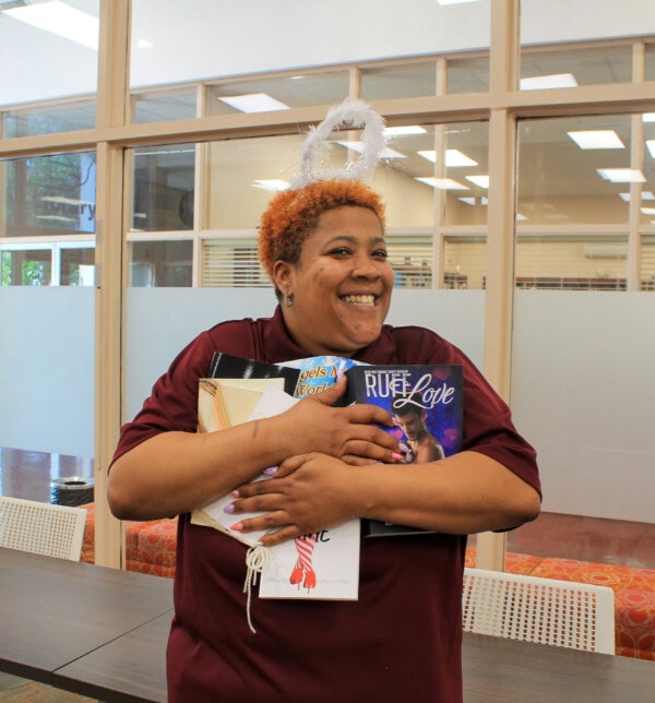 A smiling person wearing a white, fluffy halo headband hugs a stack of books in a library, standing by a table with chairs and glass walls in the background.