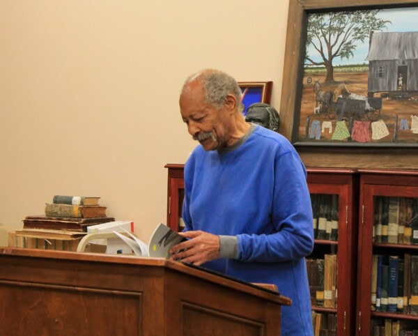 An elderly man in a blue sweater stands at a wooden podium reading a book in a library. Behind him are shelves of books and a framed painting depicting a rural scene with a house and trees.