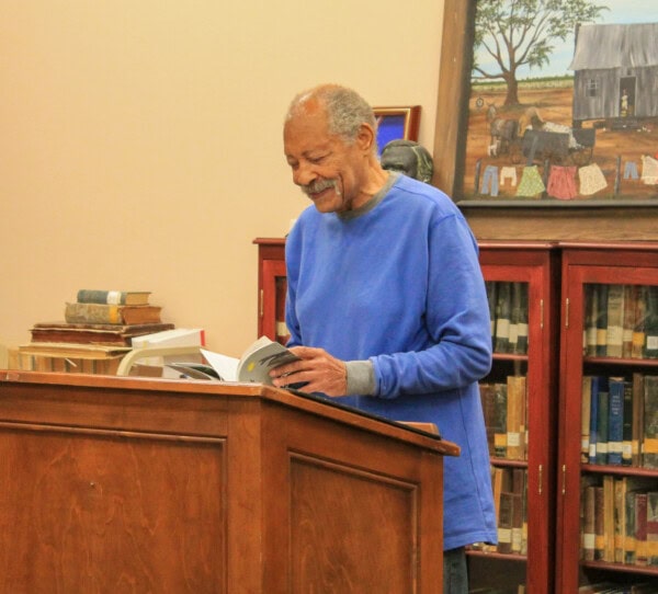An older man in a blue sweater stands at a wooden podium, smiling and reading from a book in a library. Bookshelves and a painting of a rural scene are visible in the background.