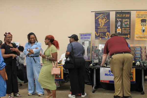 A group of people interact near tables with books, posters, and merchandise at an indoor event, with some browsing and others talking and laughing.