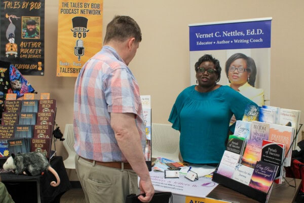 A woman stands behind a table displaying books and promotional materials, speaking to a man at a book fair. A banner identifies her as Vernet C. Nettles, Ed.D., educator, author, and writing coach.