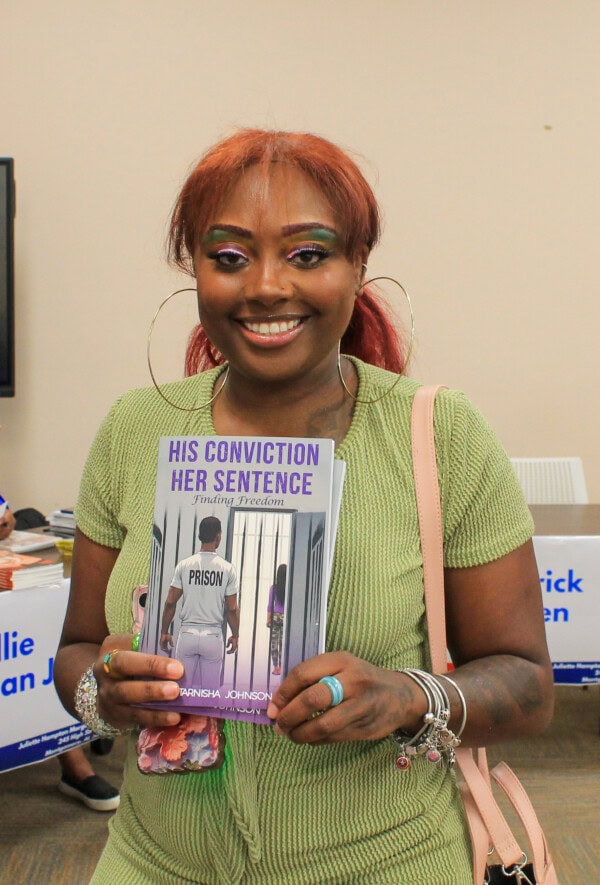A smiling woman with red hair and colorful eye makeup holds a book titled His Conviction Her Sentence: Finding Freedom in a room with tables and signs in the background.