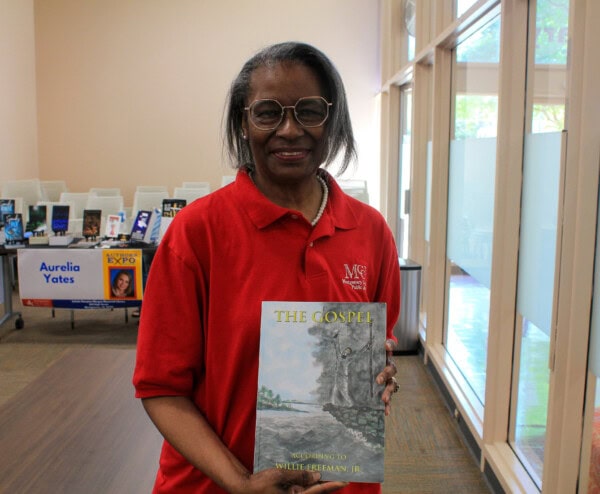 A woman wearing glasses and a red polo shirt smiles while holding a book titled The Gospel. Behind her are books displayed on tables, one with a sign that reads Aurelia Yates. Large windows let in natural light.