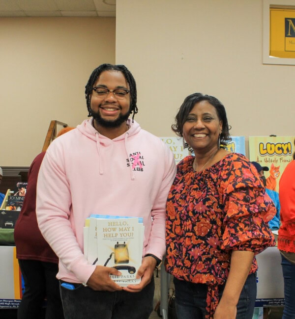 A smiling man in a light pink hoodie holds several books while standing next to a smiling woman in a red floral blouse, with a display of colorful books visible in the background.