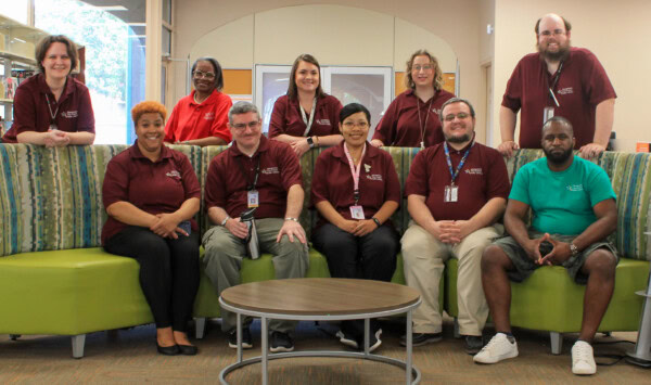 A group of ten adults, mostly wearing maroon shirts with name badges, sit and stand on green chairs in a library or office space, smiling at the camera. One person wears a green shirt and shorts.