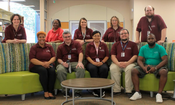 A group of ten adults, most wearing matching maroon shirts, sit and stand around a green sofa in a library or office setting, smiling at the camera.